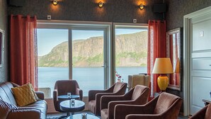 A room with a sofa, armchairs, a coffee table, and a lamp. There are curtains and a view of a body of water and hills. - Arctic Panorama Lodge (Skjervoy)