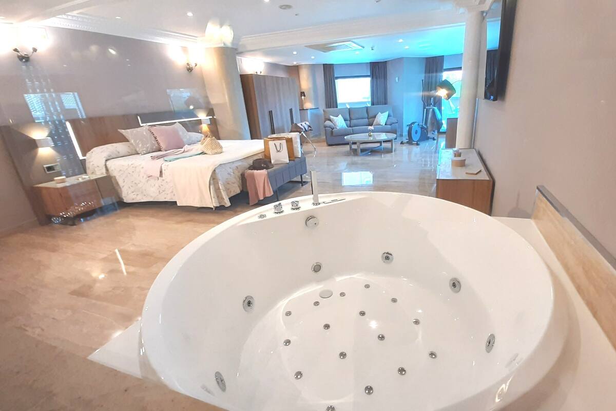 A spacious bedroom with a large bathtub, a sofa, and a dining area in the background.