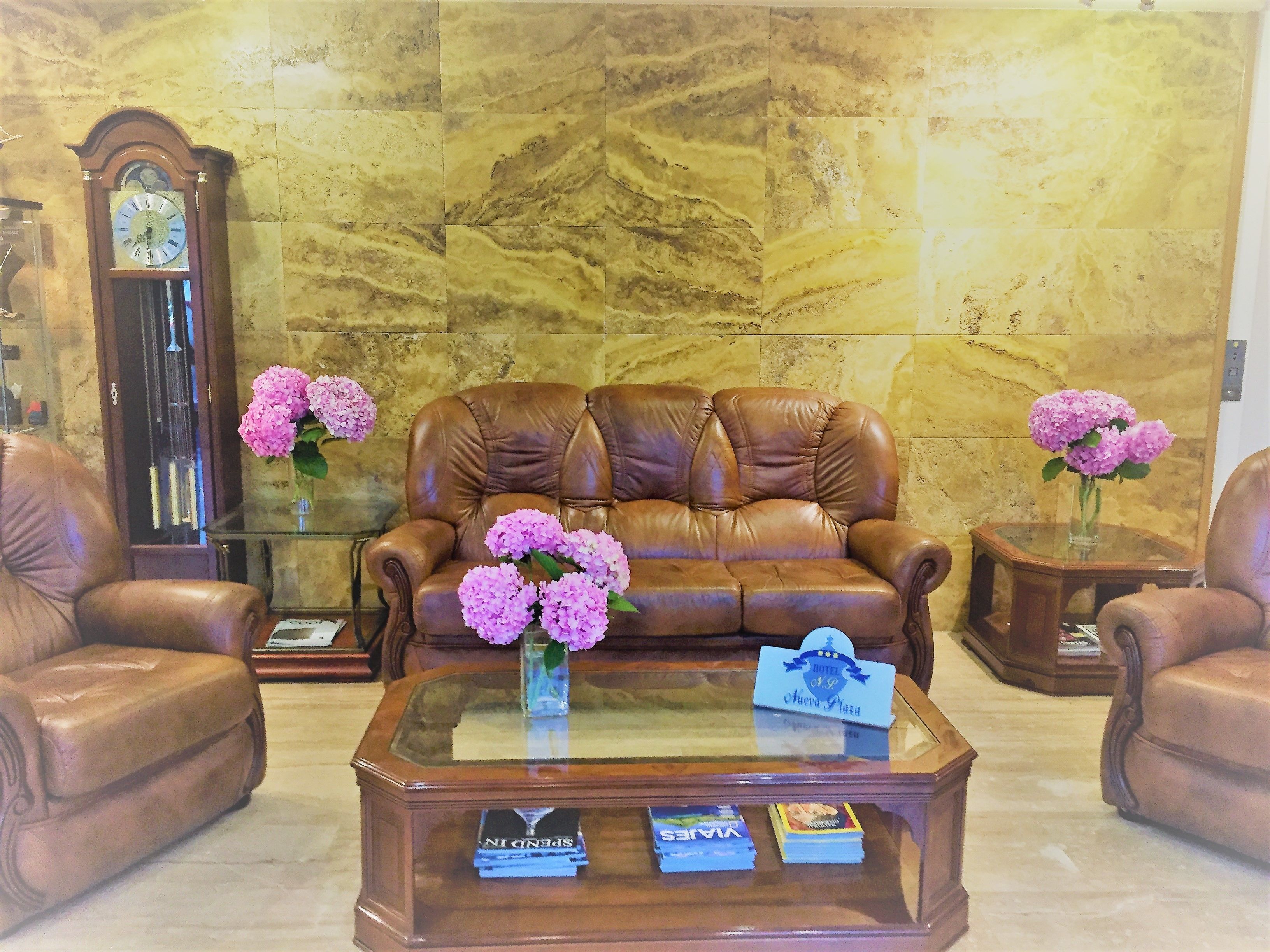 A lobby with a brown leather sofa, two side chairs, a glass coffee table with books, and a clock on the wall.