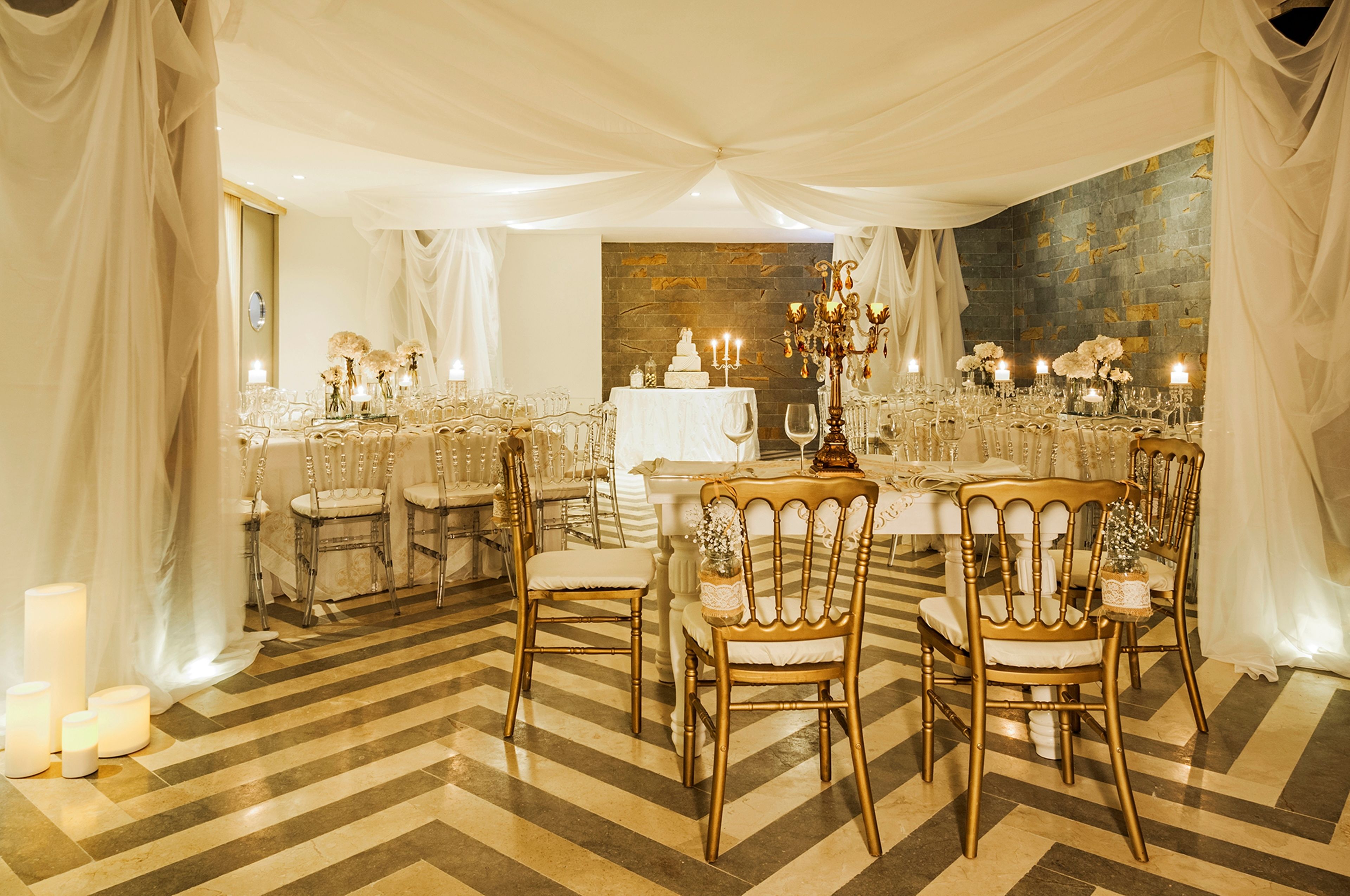 Un salón de banquetes con una larga mesa dispuesta para un evento formal, rodeada de sillas, velas y una decoración central.