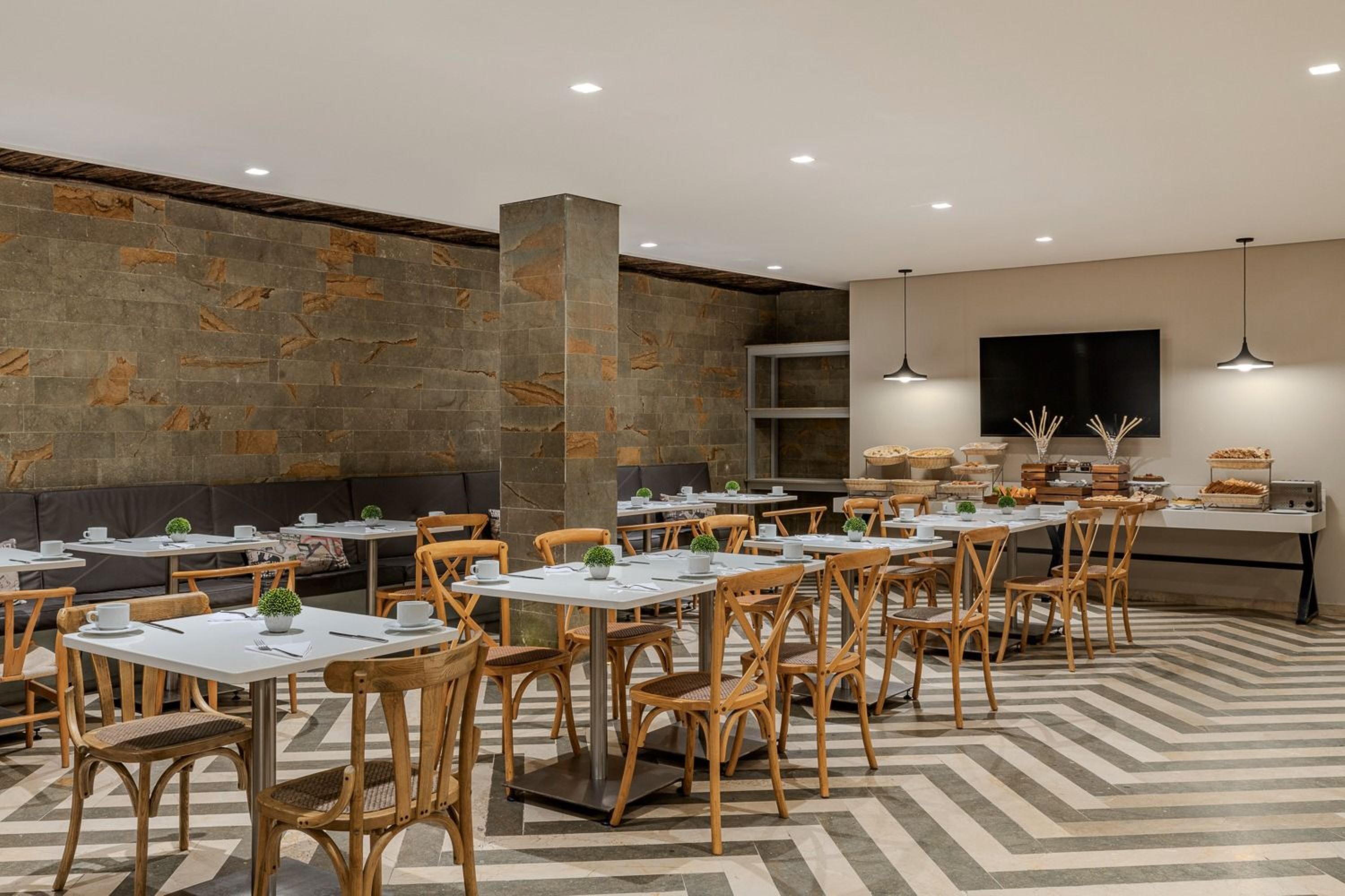 A modern restaurant with multiple tables and chairs, a stone wall, and a flat-screen TV mounted on the wall.