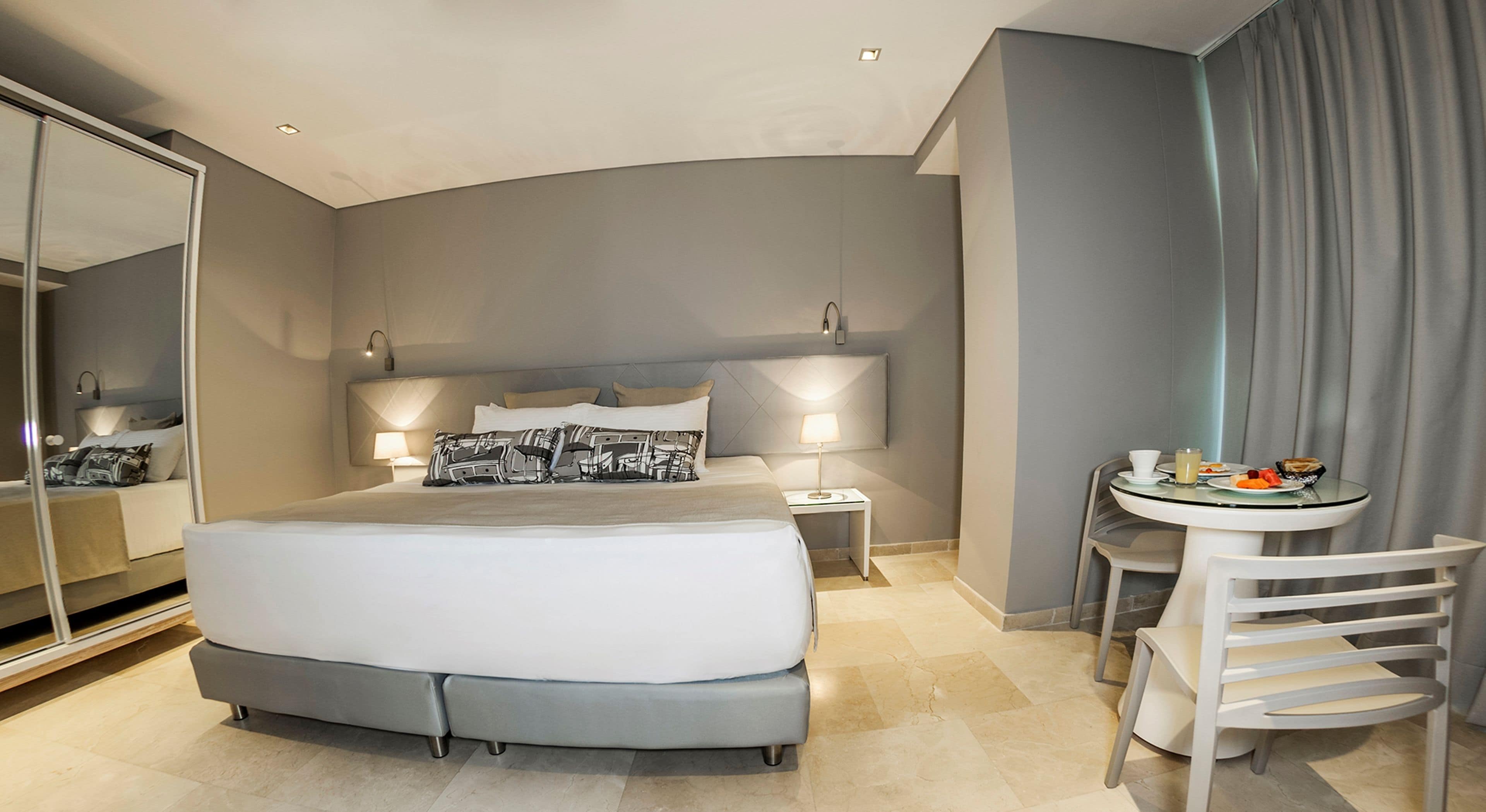 A modern hotel room with a large bed, bedside tables, a small round table with breakfast, and a mirror.