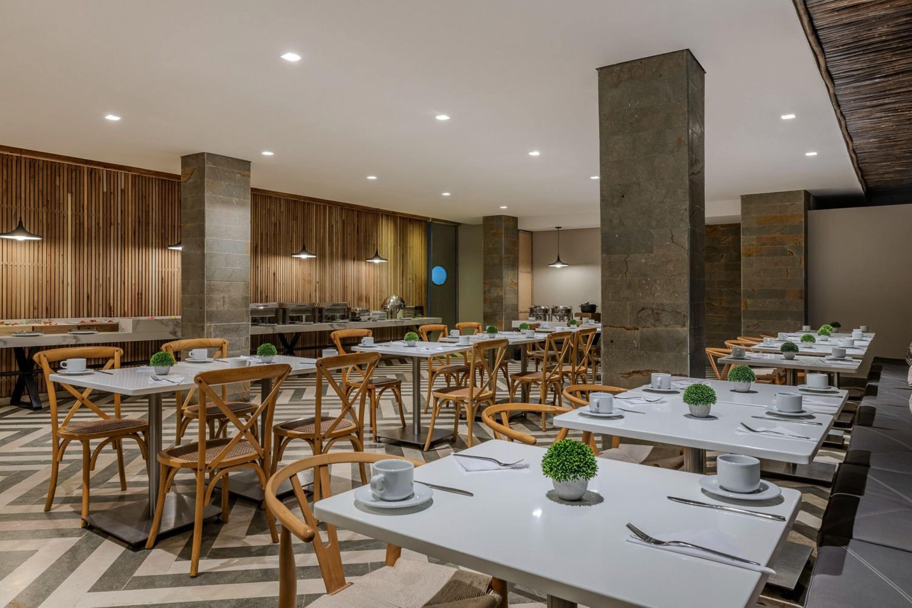 A modern dining area with wooden chairs and white tables, featuring a central column and a kitchen in the background.