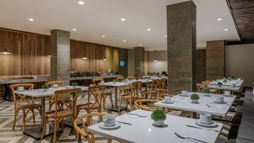 A modern dining area with wooden chairs and white tables, featuring a central column and a kitchen in the background.