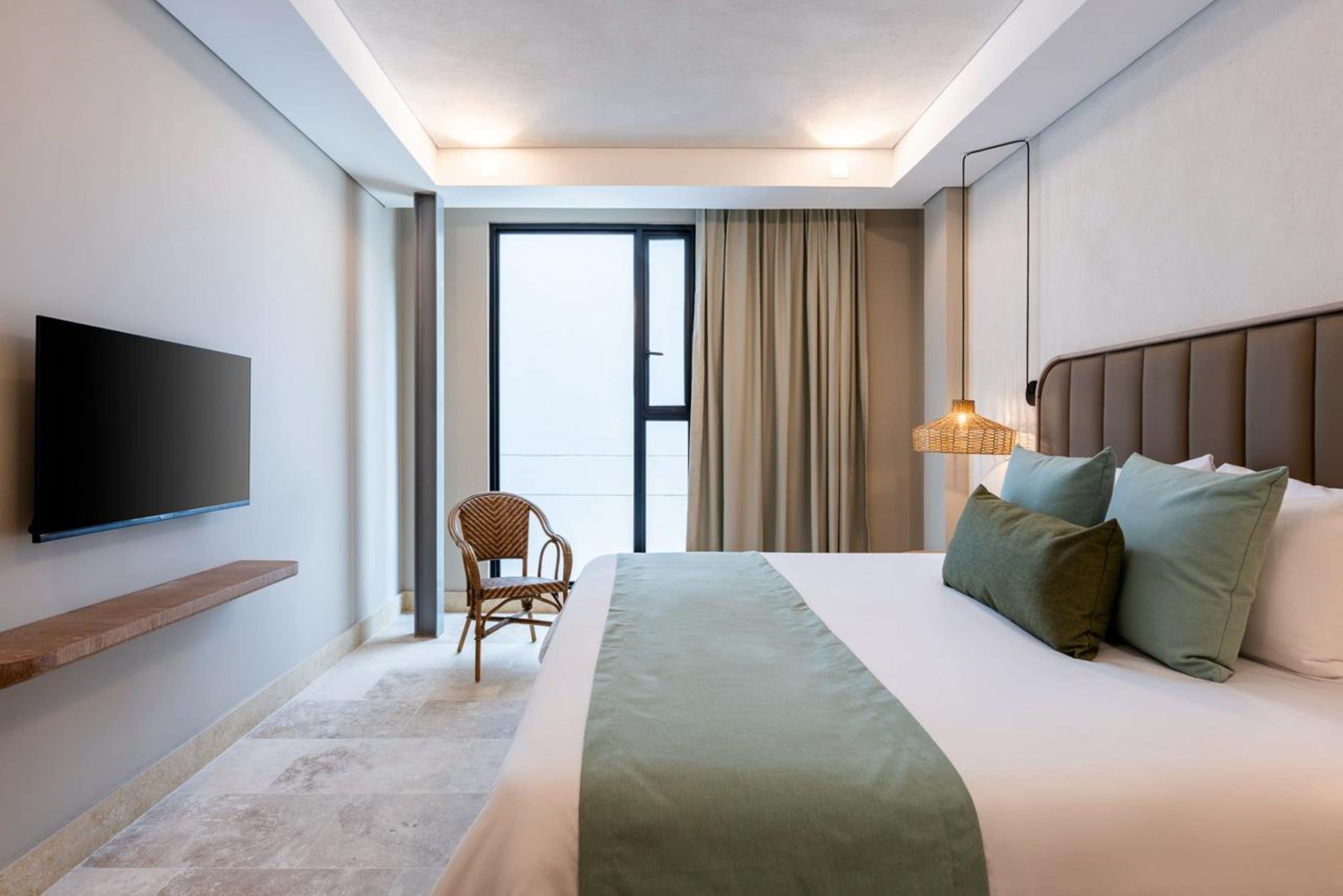 Una habitación de hotel con cama, televisión montada en la pared, una silla y ventana con cortinas.