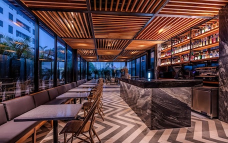 A modern bar area with a long counter, seating along the wall, and a large window offering a view of the city.