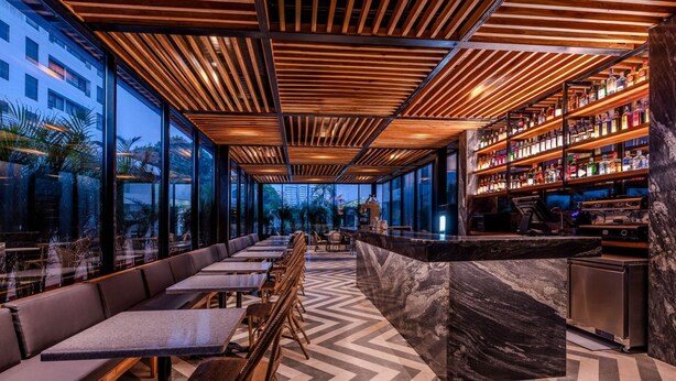 A modern bar area with a long counter, seating along the wall, and a large window offering a view of the city. - Hotel OZ Grand (Cartagena)