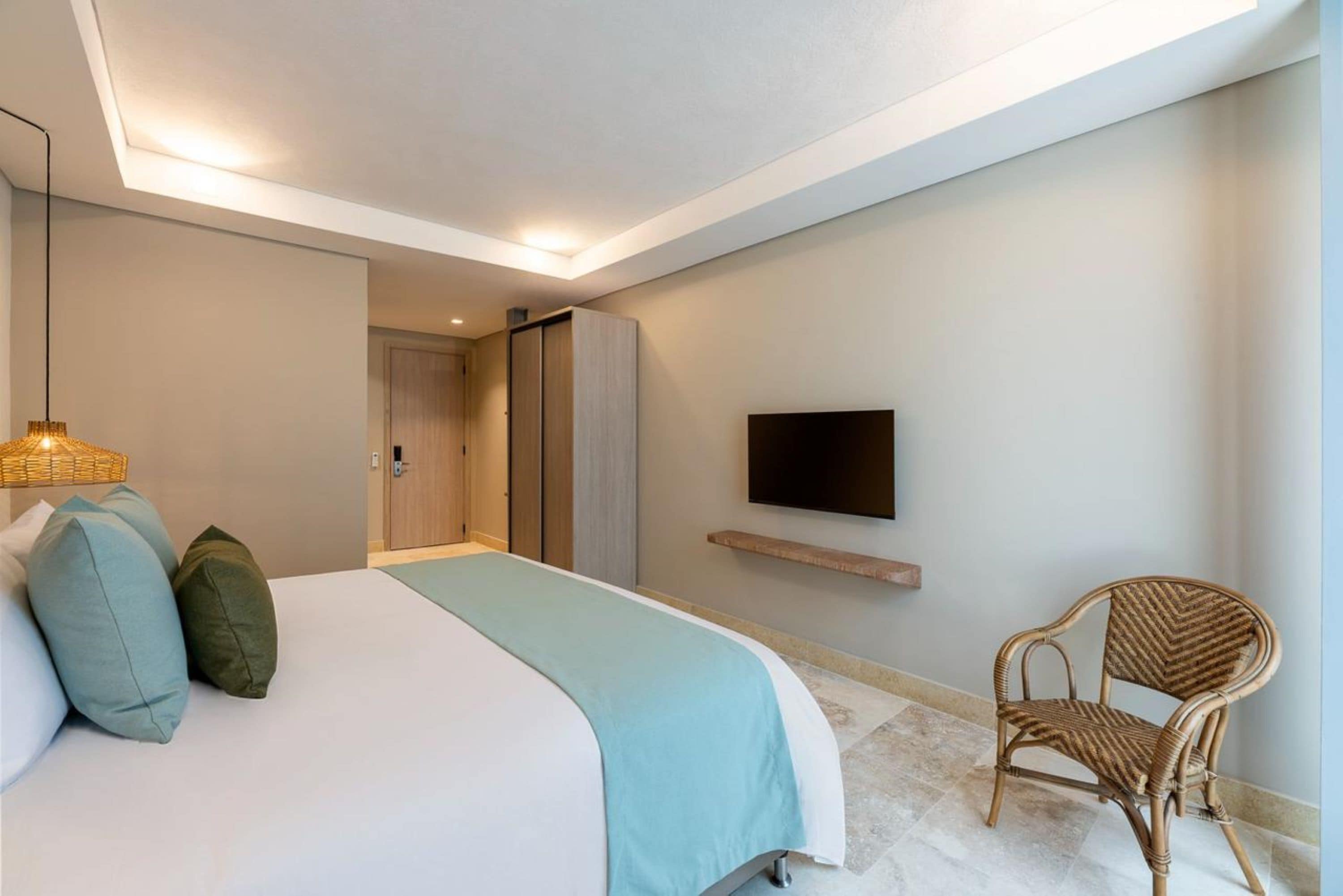 A modern bedroom with a large bed, a flat-screen TV mounted on the wall, a wicker chair, and a small wooden shelf.