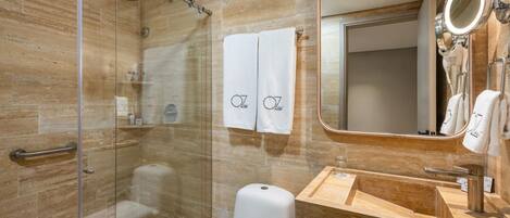 A modern bathroom with a glass-enclosed shower, a white toilet, a wooden vanity with a sink, and a large mirror with a round light.