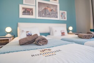 Habitación de hotel con dos camas, cada una con una colcha azul y un almohadón con el nombre del hotel. En la pared hay cuadros enmarcados y una lámpara a cada lado de las camas.