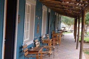 Un área de estar al aire libre techada con mesas y bancos de madera, un sendero pavimentado de piedra y un edificio con paredes azules y adornos blancos.