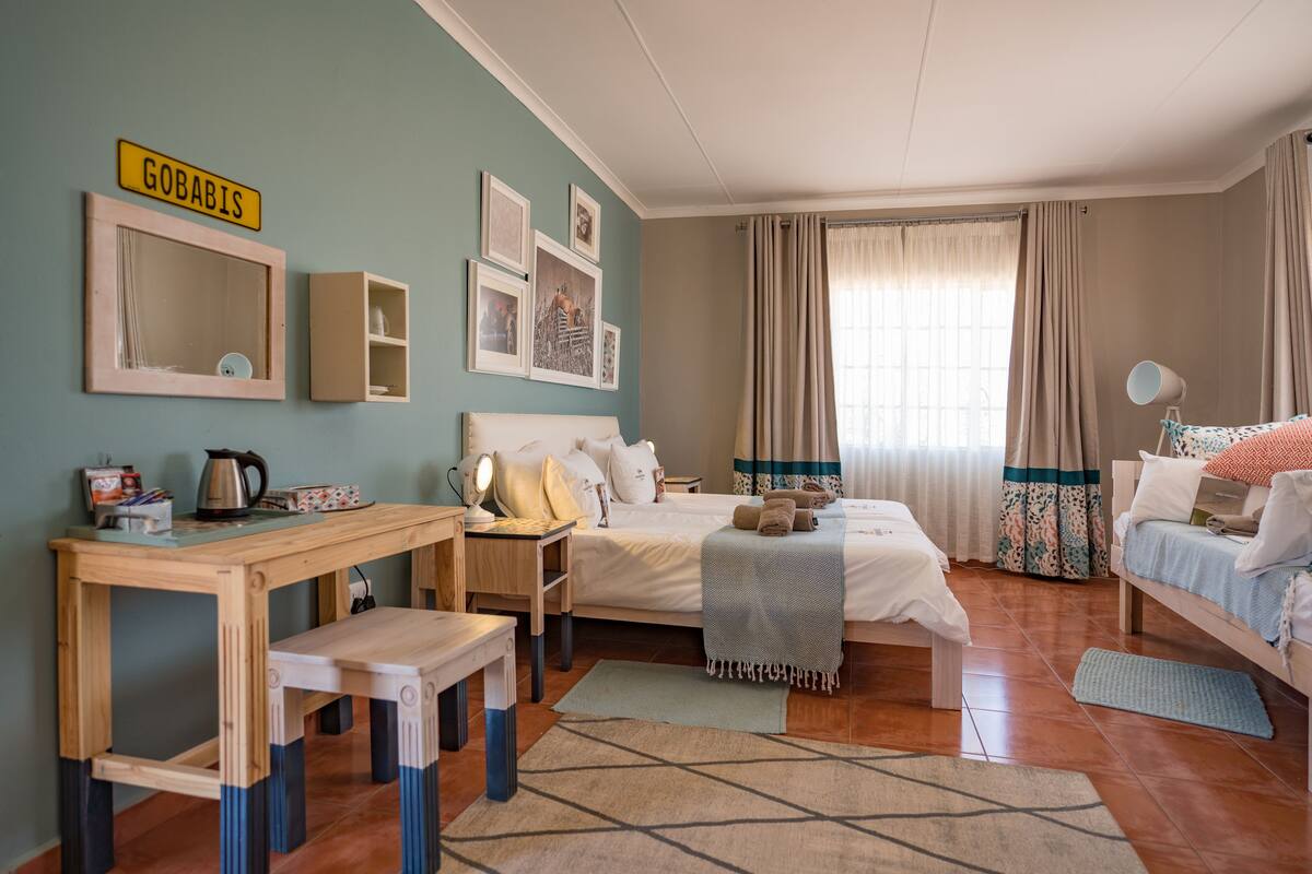 Un dormitorio con cama, mesita de noche, lámpara y escritorio con silla. Hay cortinas, una ventana y una estantería con fotografías.