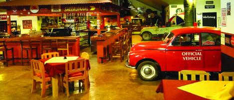 Un restaurant à la décoration d’époque, avec des meubles en bois et une voiture rouge ancienne, portant l’inscription « VÉHICULE OFFICIEL », garée à l’intérieur.