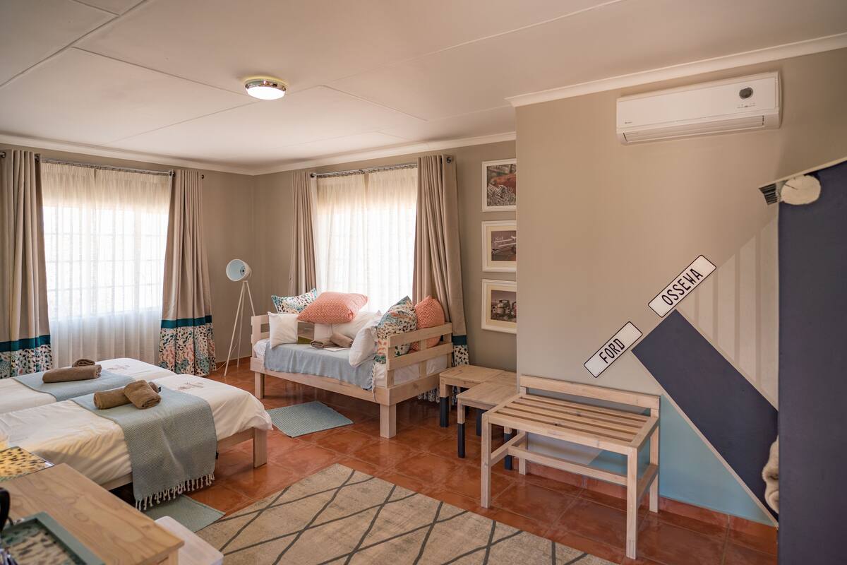 Un dormitorio acogedor con dos camas, una mesita de noche, un escritorio pequeño y una silla. Se aprecia una ventana con cortinas, un ventilador de techo y un aire acondicionado de pared.