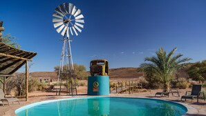Zwembad met een windmolen, omringd door woestijnlandschap en tuinmeubilair.