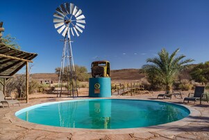 Una piscina con un molino de viento, rodeada de paisaje desértico y mobiliario exterior.