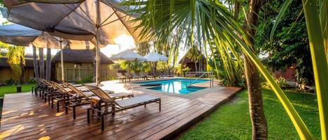 Una terraza de madera con sillones de descanso y sombrillas, una piscina y follaje tropical.