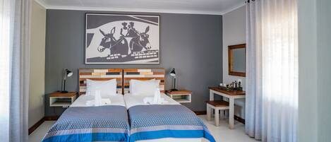 Una cama doble con colcha estampada, mesitas de noche con lámparas, cabecera con diseño de paneles de madera, un cuadro de un burro en la pared y un ventanal con cortinas traslúcidas.