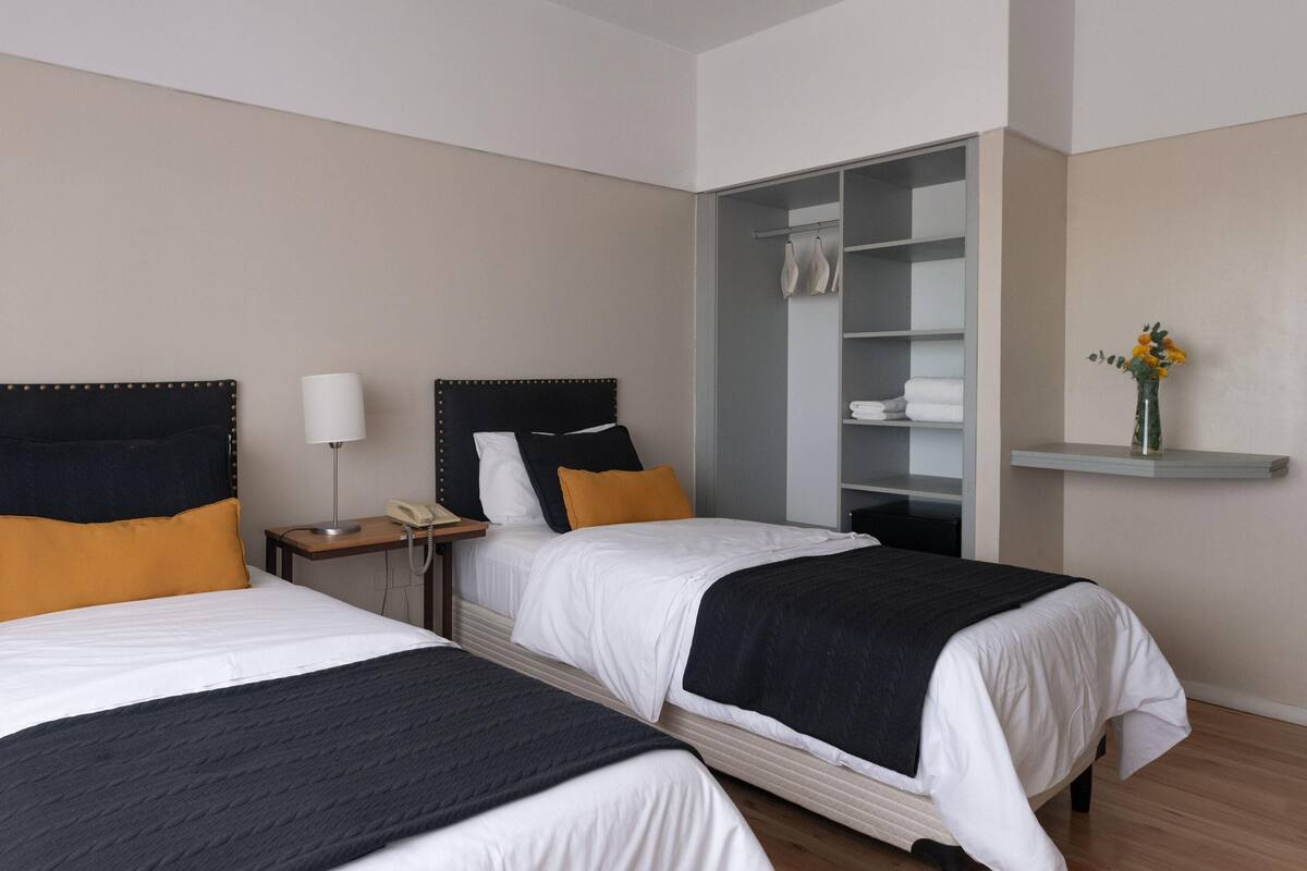 Une chambre d’hôtel avec deux lits, une armoire, une table de chevet avec une lampe et une étagère avec des fleurs.