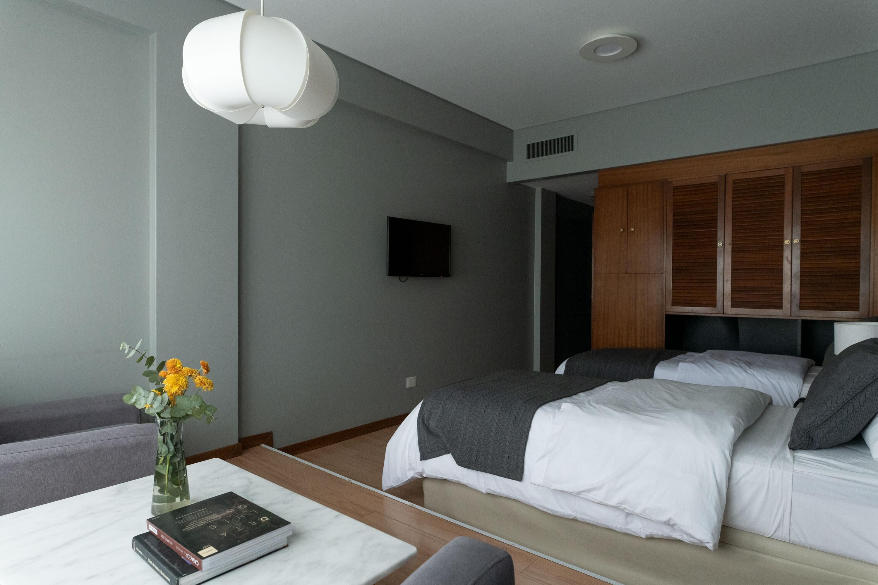 A modern bedroom with a bed, a flat-screen TV, a wooden wardrobe, and a vase with flowers on a nightstand.