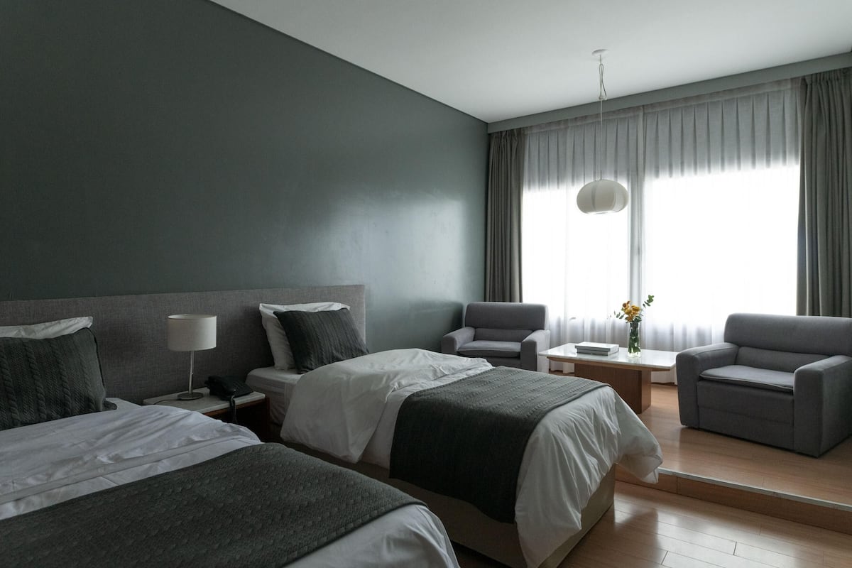 Une chambre d’hôtel avec deux lits, un bureau, une chaise et une grande fenêtre avec des rideaux.