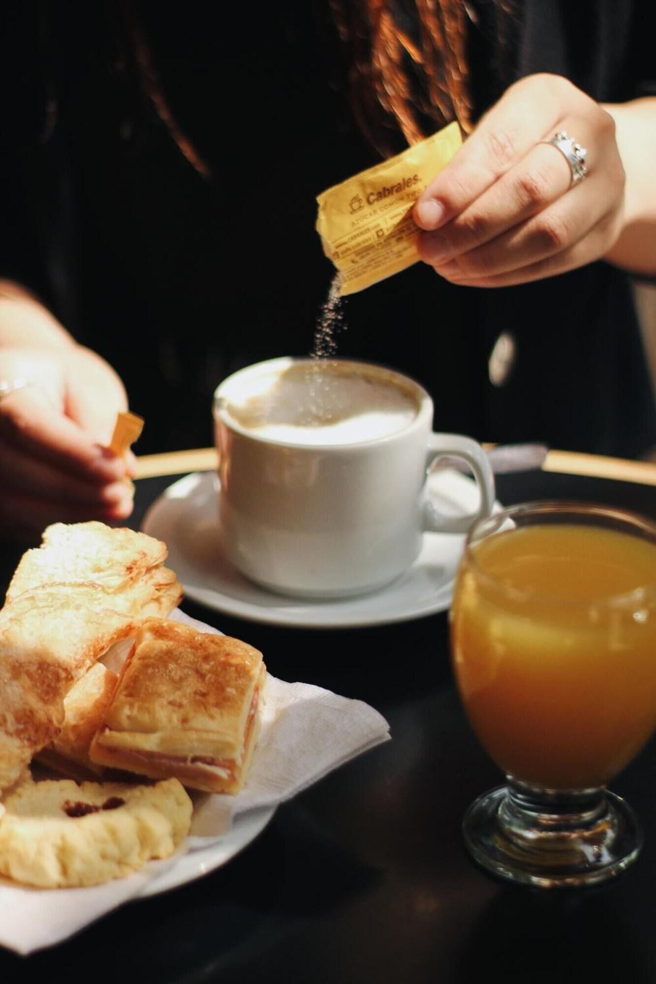 A person pouring sugar into a cup of coffee, a plate of pastries, and a glass of tea.