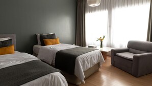 A hotel room with two beds, a grey armchair, a small table with a vase, and a large window with curtains. - Aguada Hotel (Trelew)