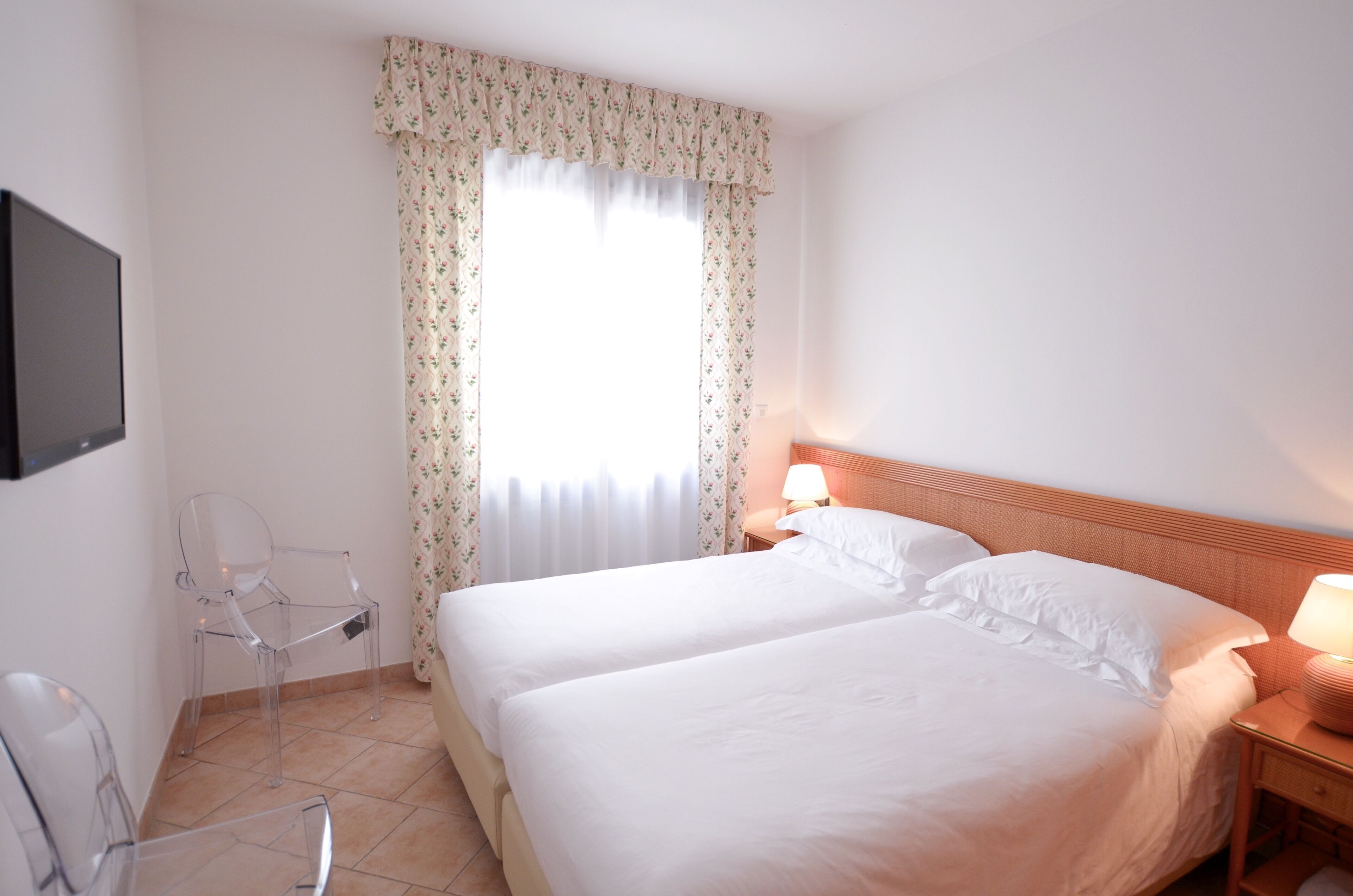 Habitación de hotel con dos camas, una silla, una mesita de noche con lámpara y un televisor montado en la pared.