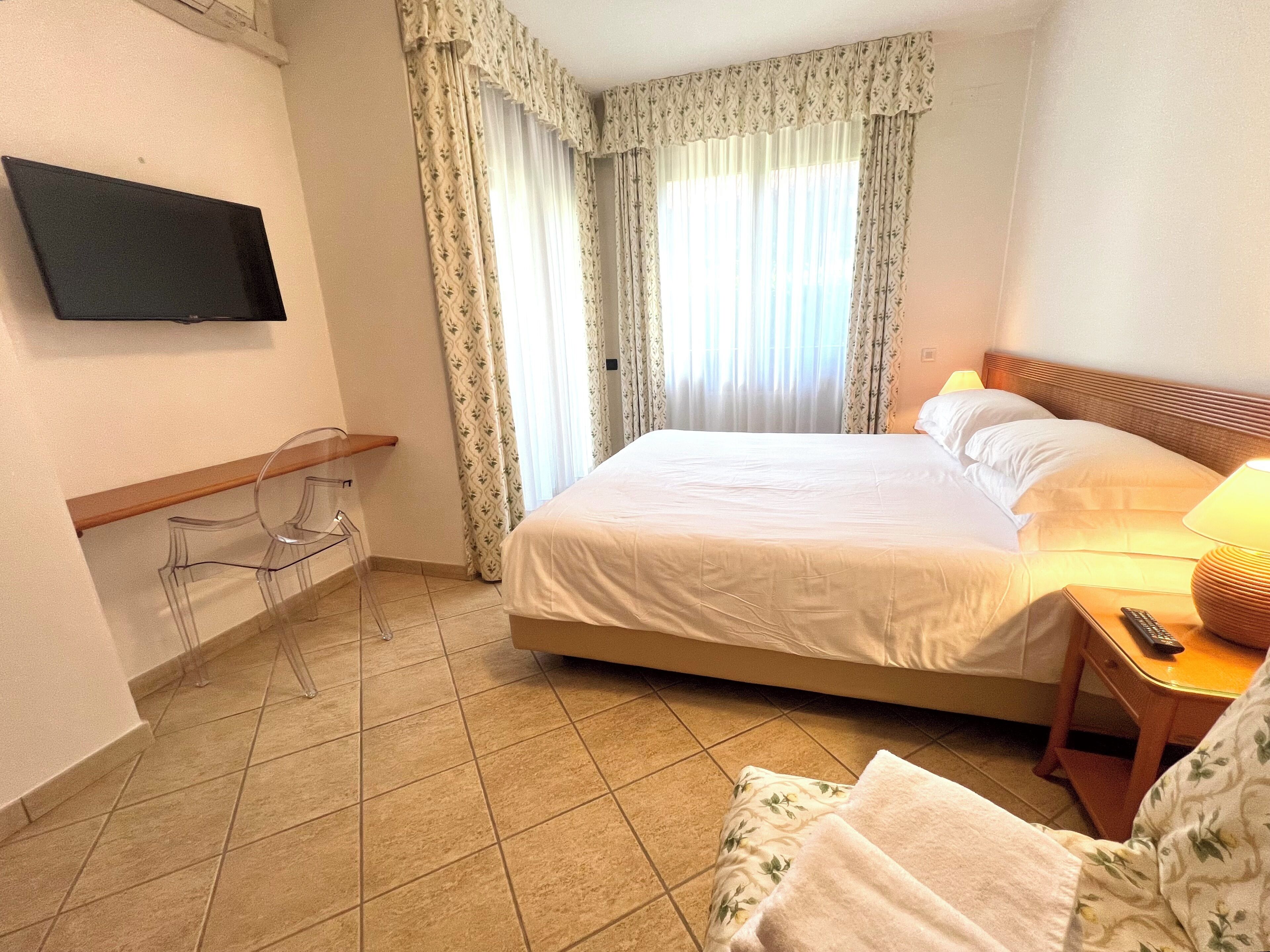 A hotel room with a bed, a TV mounted on the wall, a bedside table with a lamp, and a chair.