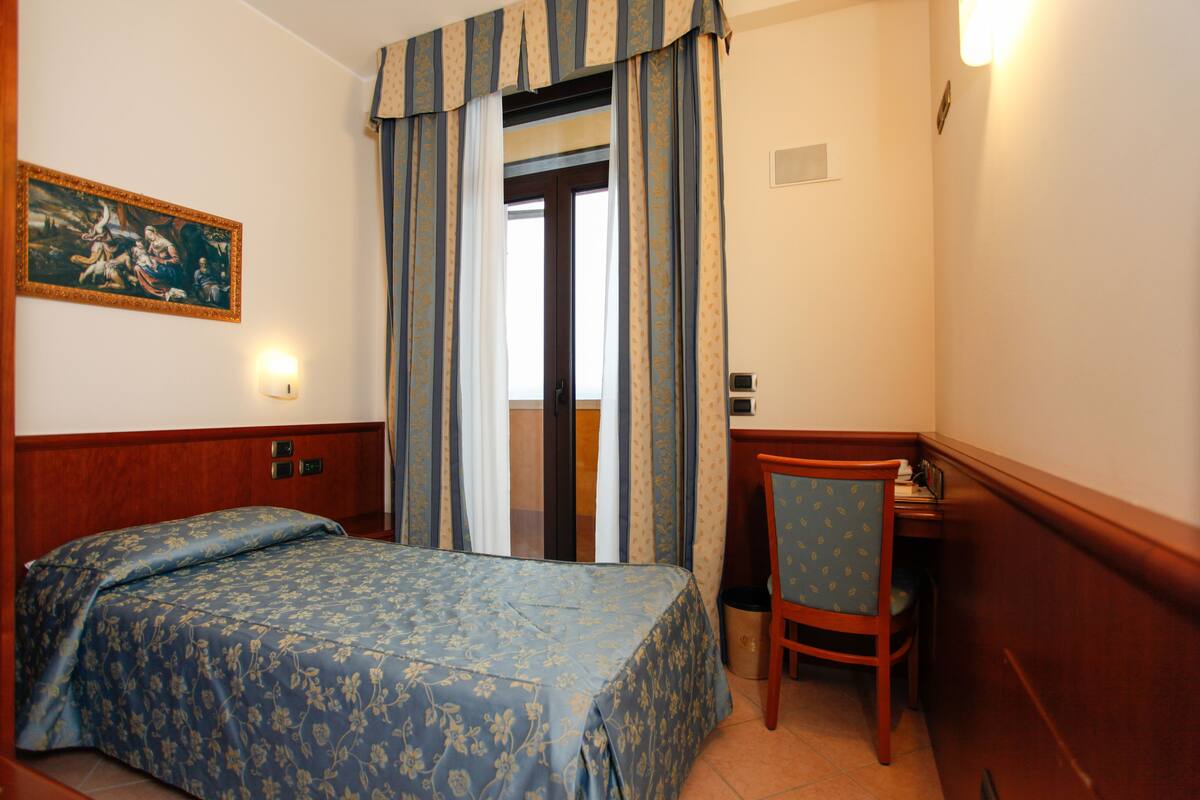 A hotel room with a bed, a desk, and a chair. There is a painting on the wall and a door with curtains.