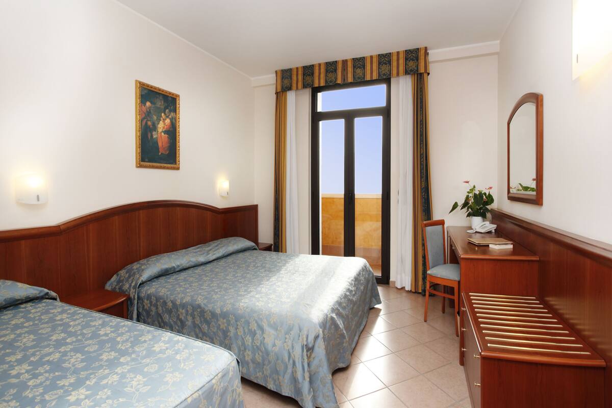 A hotel room with two beds, a desk with a chair, a window with curtains, a painting on the wall, and a mirror.