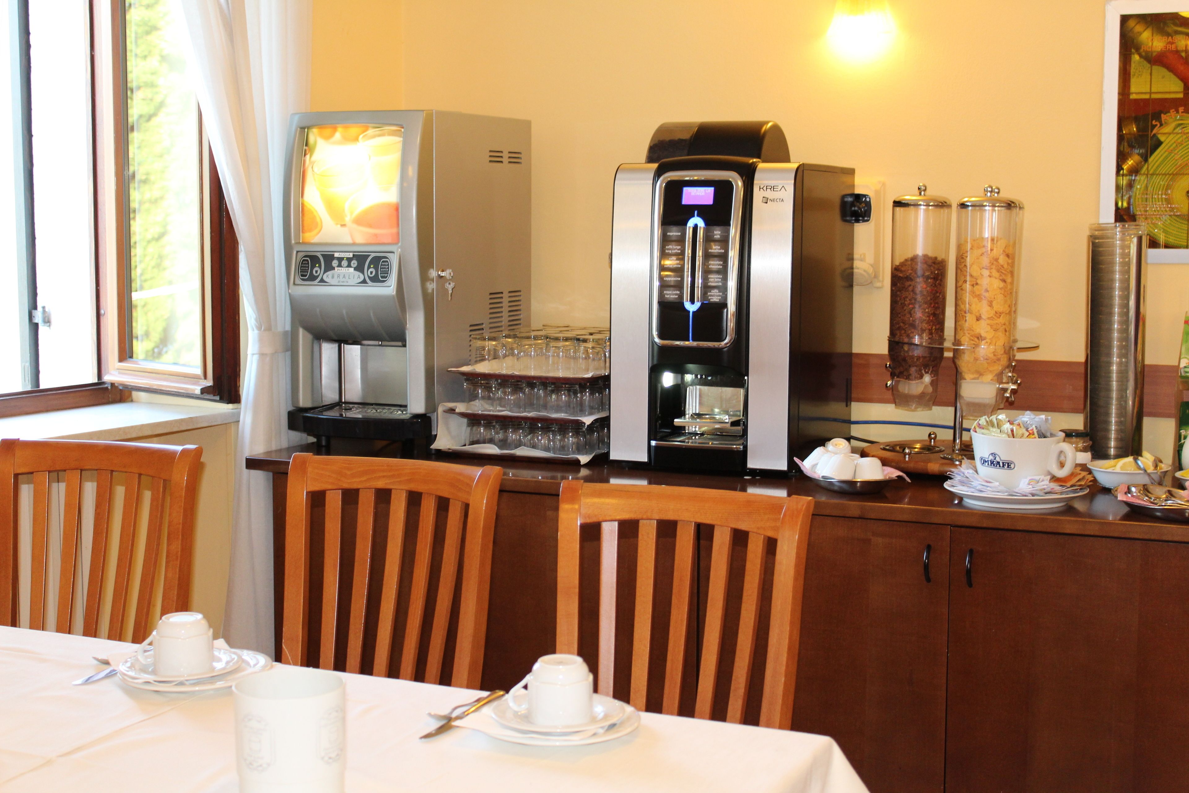 Uma área de café da manhã com cafeteira, um expositor de vidro e uma mesa de jantar posta com xícaras e pireses.