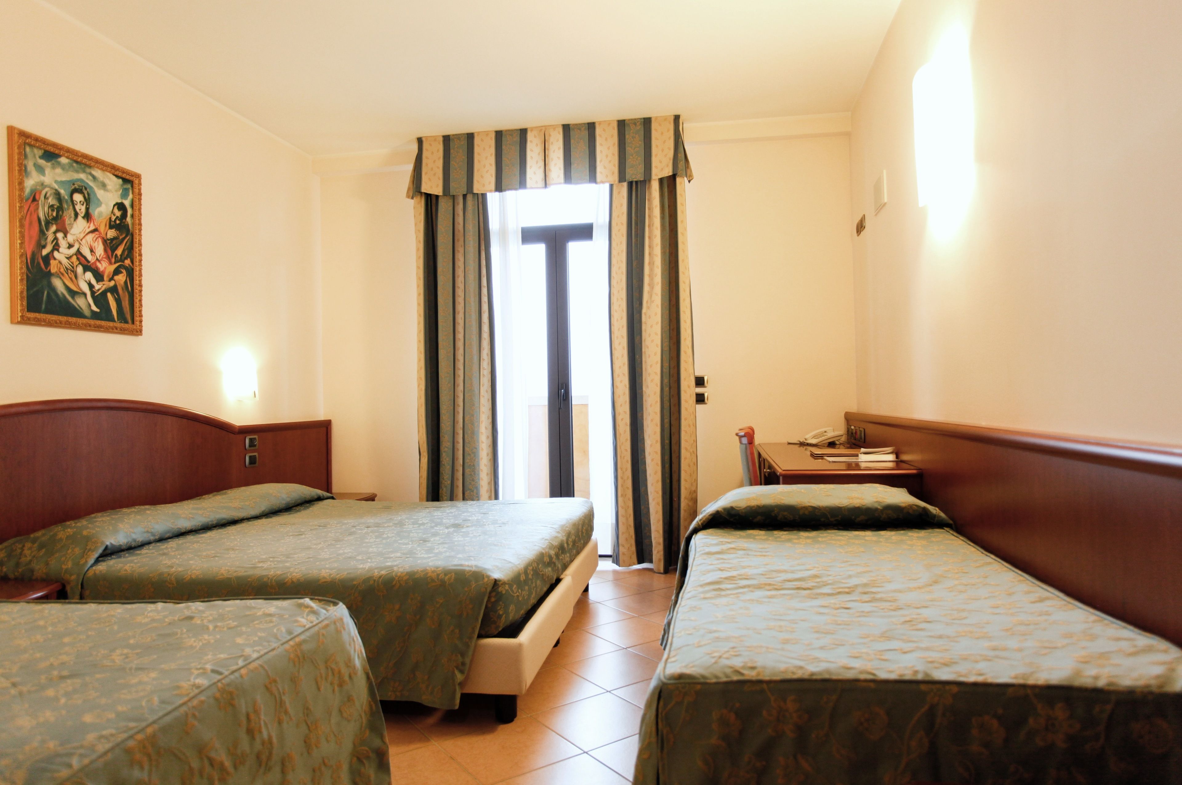 Ein Hotelzimmer mit zwei Betten, einem Bild an der Wand, einem Fenster mit Vorhängen und einem Nachttisch mit Lampe.