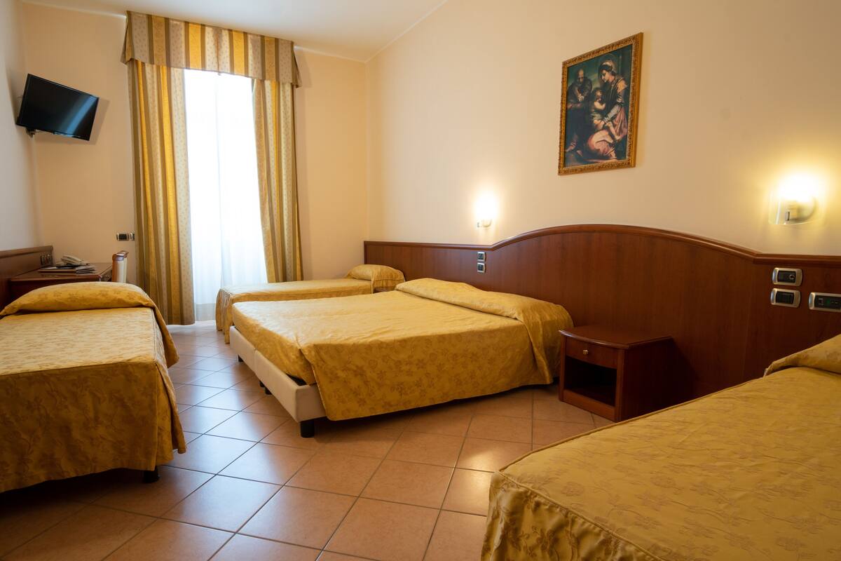 A hotel room with three beds, a television mounted on the wall, a painting, and a nightstand with a lamp.
