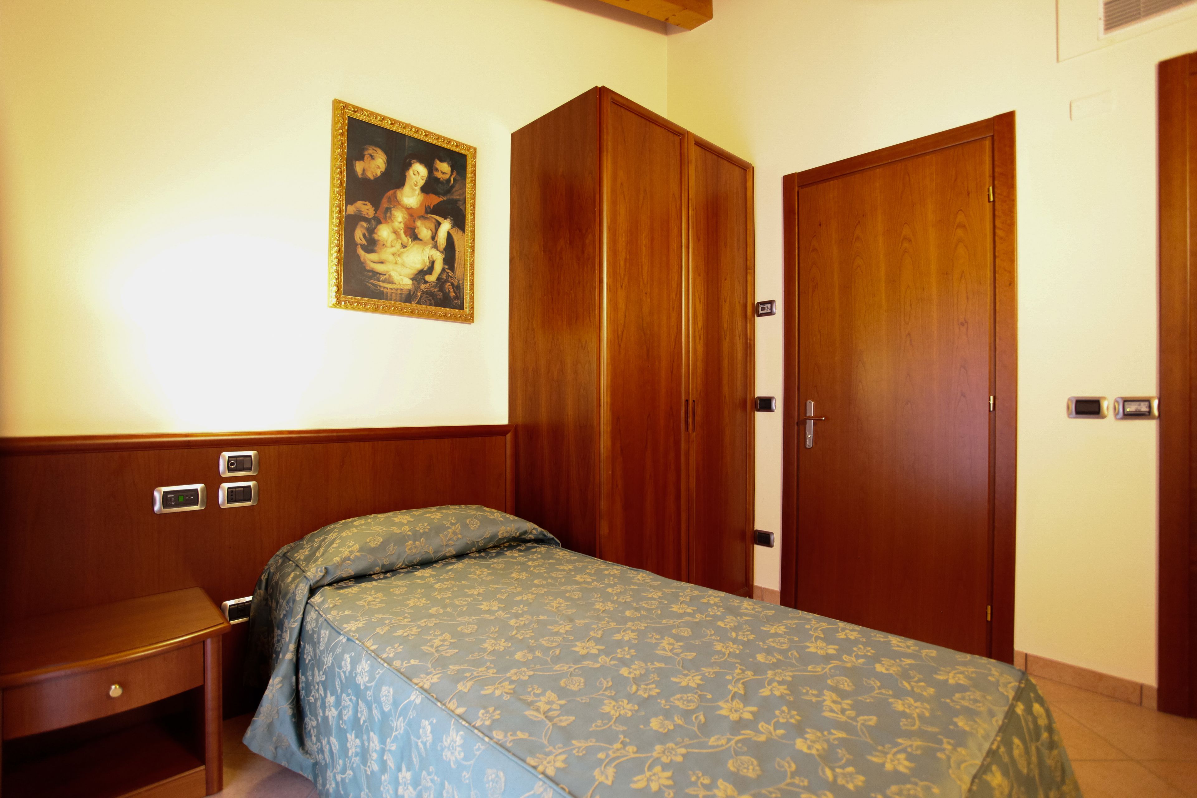 A hotel room with a bed, a wooden wardrobe, a nightstand, and a painting on the wall.