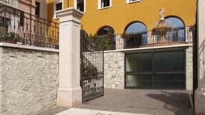 Una entrada de piedra con un portón de metal, un edificio amarillo con ventanales arqueados y una fuente.