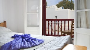 A bedroom with a bed, a wooden nightstand, a window with a view of the sky and buildings, and a balcony with a chair and table.