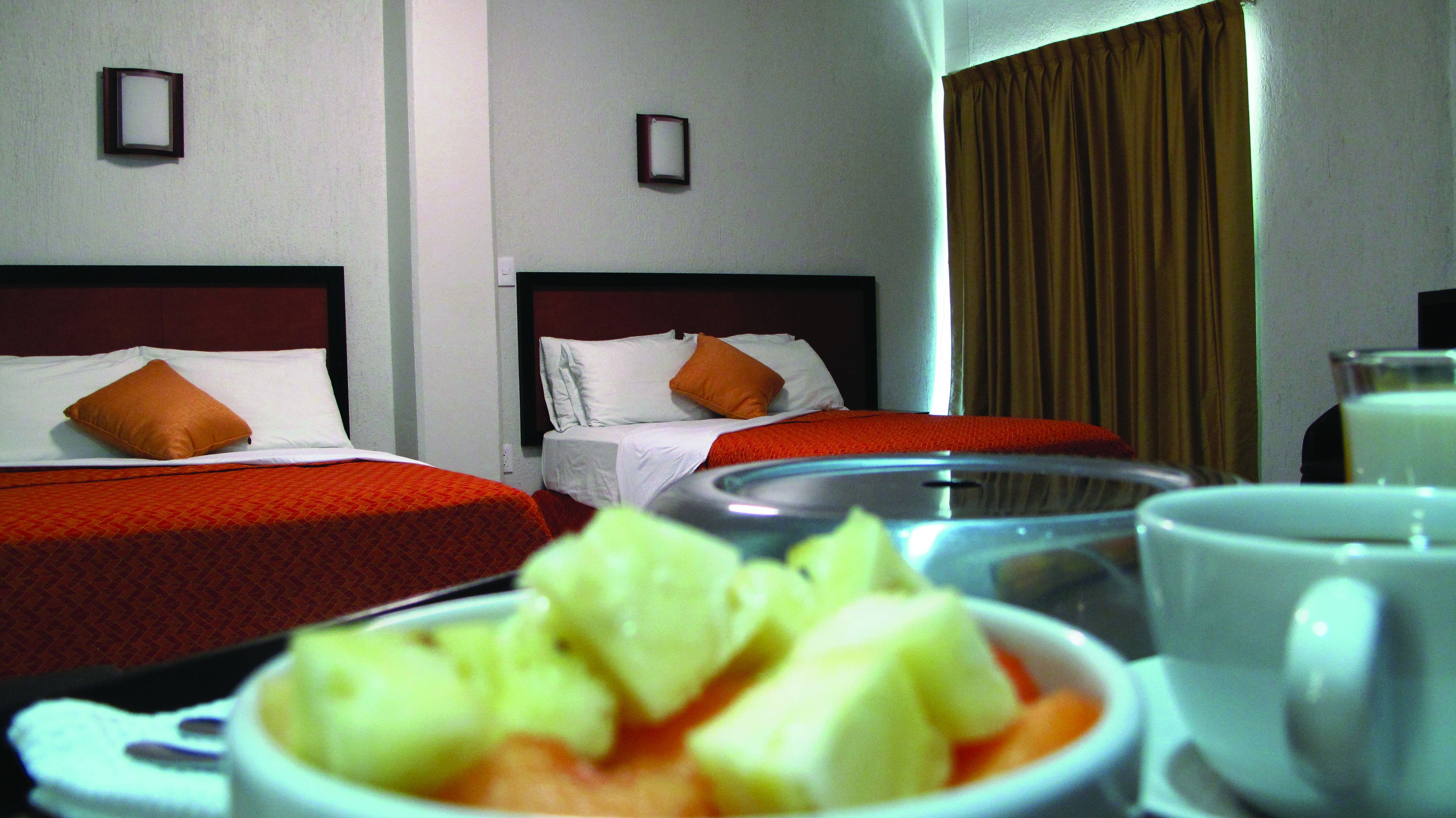 Dos camas individuales con ropa de cama roja y almohadas naranjas, una mesa de comedor con un bol de fruta y una taza, y una ventana con cortinas.