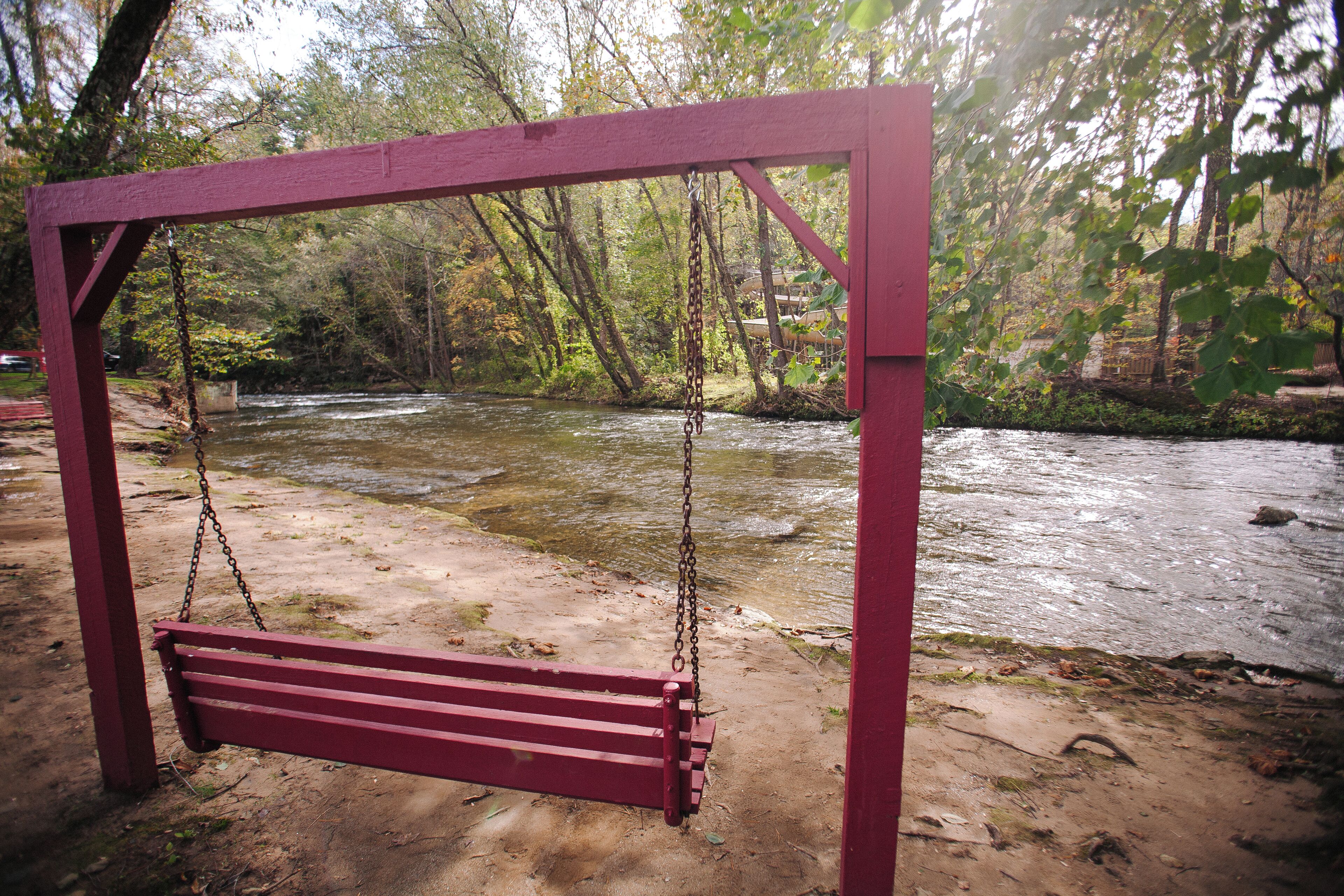 Un columpio rojo junto a un río, con un sendero de arena y árboles al fondo.