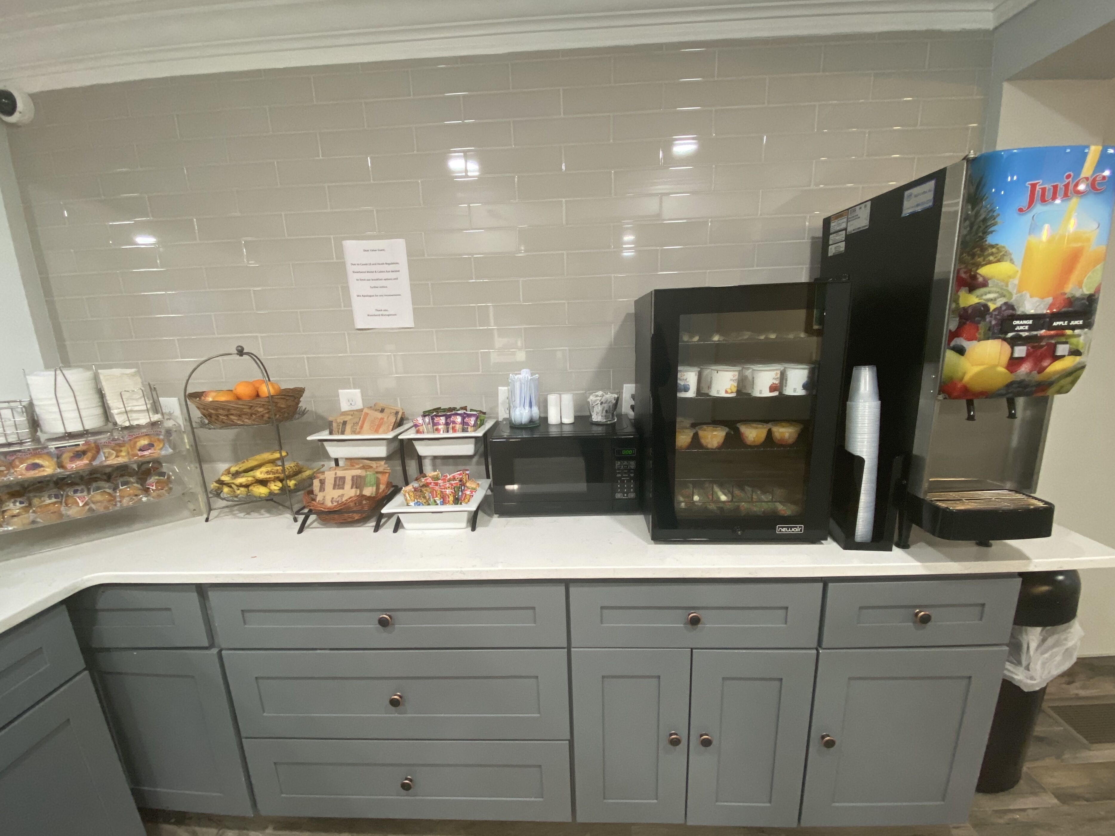 A kitchen area with a refrigerator, microwave, juice dispenser, and various food items on the counter.