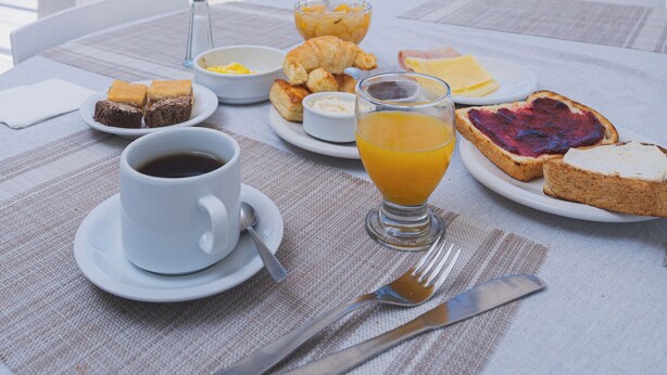 A breakfast table with croissants, toast, and orange juice, overlooking a beach with parked cars and a clear sky.