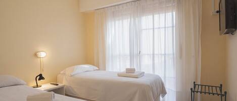 Habitación de hotel con dos camas, una mesita de noche con lámpara, un televisor montado en la pared y una ventana con cortinas traslúcidas.