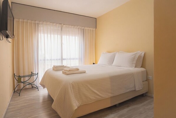 A bedroom with a bed, a window with curtains, a bedside table, and a wall-mounted TV.