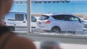 A person looking out a window at a pier with parked cars and a boat in the distance.
