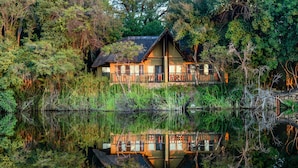 A wooden house with a balcony, surrounded by dense vegetation and a calm body of water reflecting the structure.