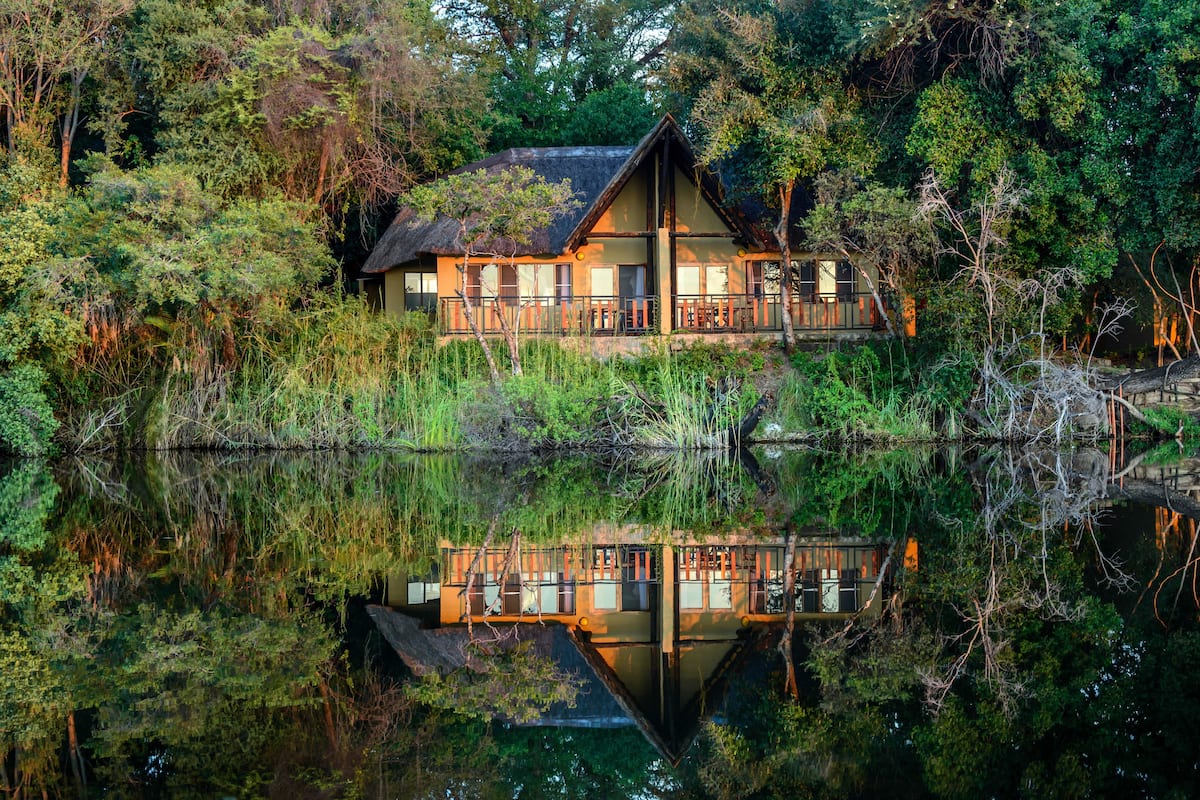 A wooden house with a balcony, surrounded by dense vegetation and a calm body of water reflecting the structure.