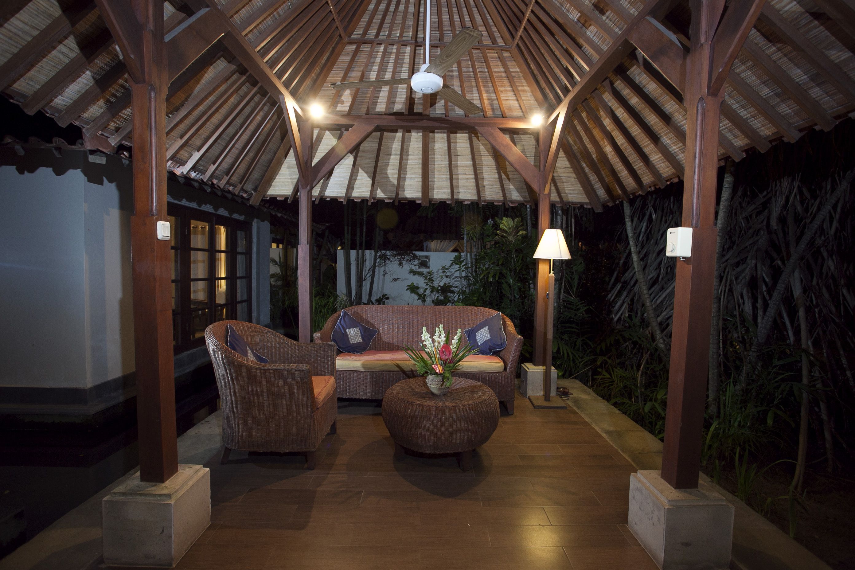 A covered outdoor seating area with wicker furniture, a central round table with a floral arrangement, and a ceiling fan.