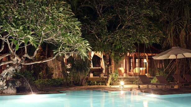 Zona de piscina con sillas de descanso y una sombrilla, rodeada de vegetación tropical.
