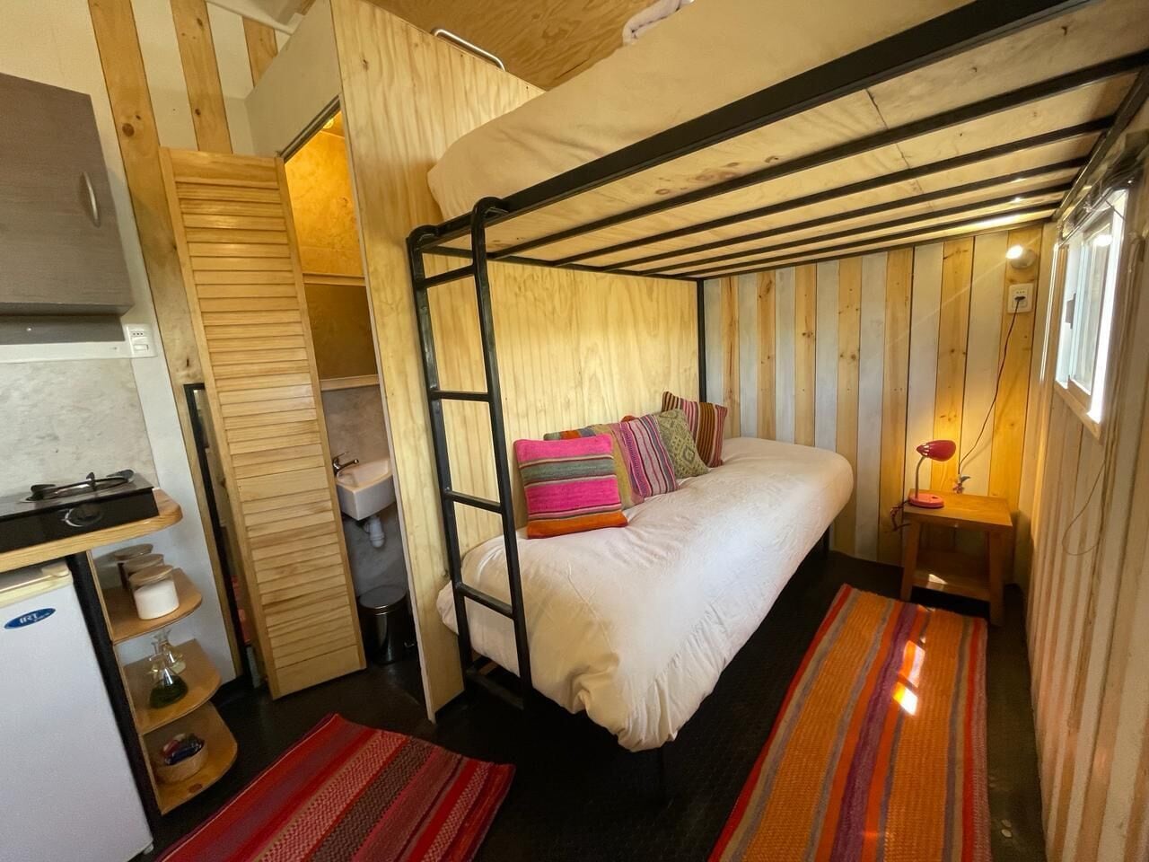 A compact, wooden cabin-style room with a bunk bed, a small kitchenette, and a single bedside table with a lamp.