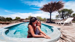 Un jacuzzi con dos personas, rodeado de madera y sillas de descanso, con un cielo azul despejado al fondo.